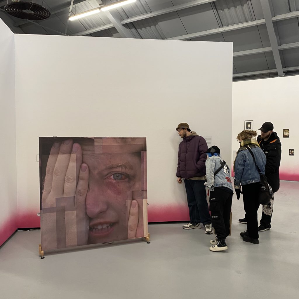 Studenter ser på samtidskunst i et gallerilokale. Kunstverket som er fokus i bildet viser nærbilde av et mannsansikt, han holder en hånd over det ene øyet og er rød rundt det andre øyet, som om han gråter.