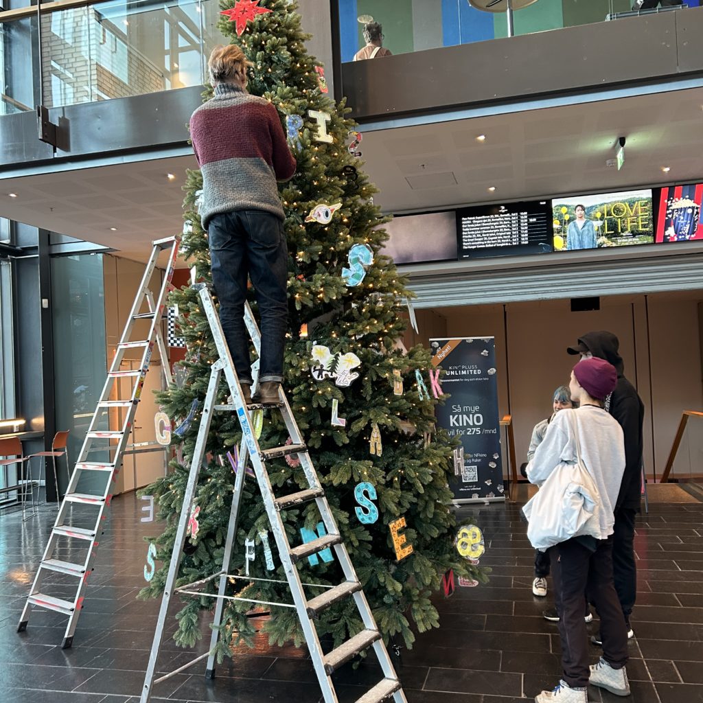 Studenter i ferd med å pynte et høyt juletre i fojeen til Asker kulturhus. Treet har lys og er pyntet med laminerte plastbokstaver i ulike farger og fasonger. En mannlig lærer står øverst i en gardintrapp og fester en av bokstavene. Tre elever står på gulvet og følger med.